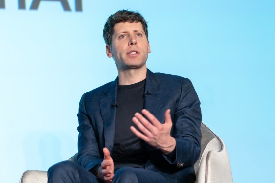 Man in a dark suit sitting and speaking with a light blue wall in the background