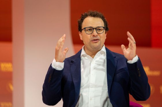 Person wearing glasses, white shirt, and dark blue blazer speaking with hands raised against blurred red and orange background