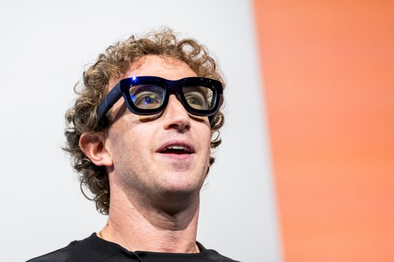 Person wearing large, black-framed glasses looking upward with neutral expression against light grey and reddish-orange background