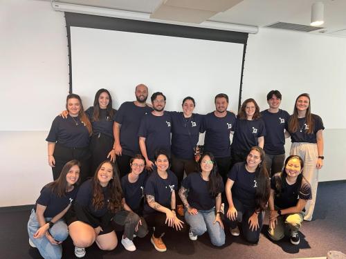 Group of people wearing matching dark blue shirts with logo, posing in conference room with projection screen
