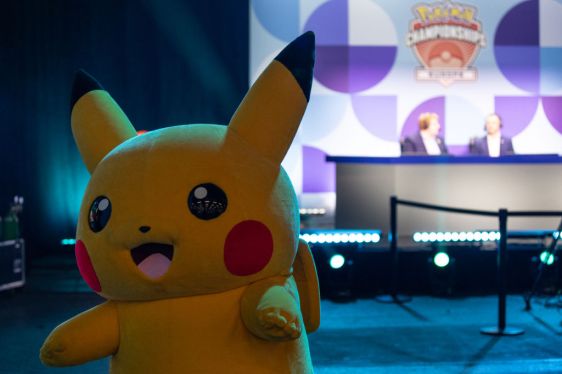 Large Pikachu costume at Pokémon Championship event with two people at a desk on a stage in the background