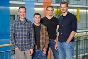 Four men standing in a modern indoor space with colorful vertical panels in the background, representing a tech or startup environment