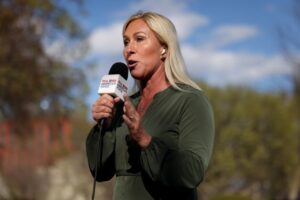 Person with long blonde hair speaking into a microphone labeled 'Real America's Voice' outdoors, with trees and a blue sky in the background, related to the controversy involving Marjorie Taylor Greene and Grok.