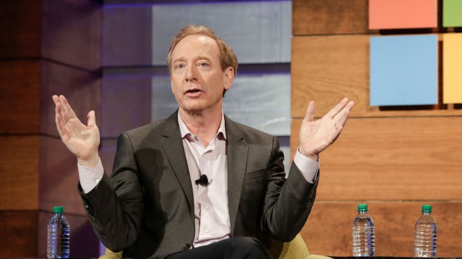 Person in suit gesturing with hands in front of a colorful wall, with water bottles on the table