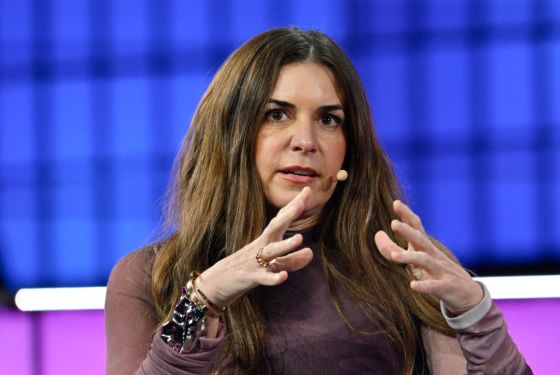 Woman speaking on stage about AI and human collaboration with a microphone, gesturing with hands, in a conference setting with purple and blue background hues.