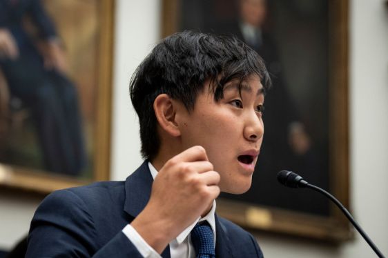 A person speaking into a microphone during a formal hearing or conference with framed portraits in the background, related to AI investment and collaboration between Meta and Scale AI.