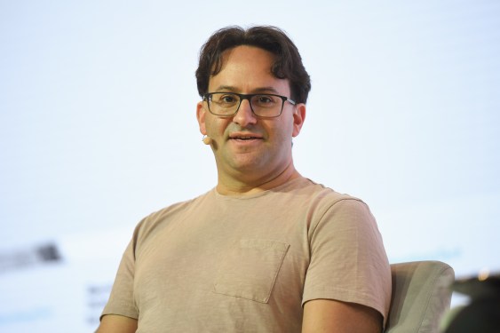 Person wearing glasses and a light-colored T-shirt speaking at an event with a microphone, blurred background
