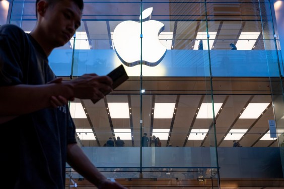 Exterior of an Apple Store with illuminated logo and glass facade, a person walking while looking at their phone, and people inside on the upper level, emphasizing Apple's focus on visual intelligence innovations.