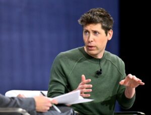 Two individuals having a conversation, one gesturing with hands and wearing a green sweater, in a discussion about OpenAI's model launch delay.
