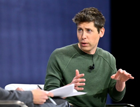 Two individuals having a conversation, one gesturing with hands and wearing a green sweater, in a discussion about OpenAI's model launch delay.