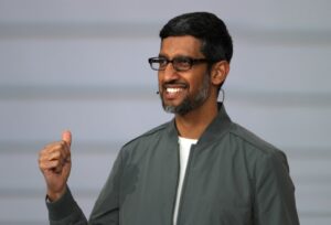 Sundar Pichai wearing glasses and a gray jacket, smiling and giving a thumbs-up during a speech or event related to growth plans